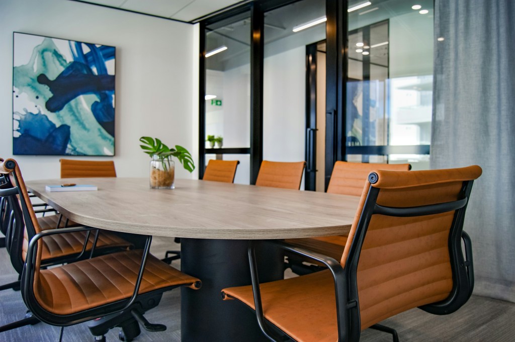 Modern boardroom with warm white walls, black trim, warm caramel coloured leather chairs, a wood table, abstract wall art, and a plant centerpiece adding comfort to a stark space